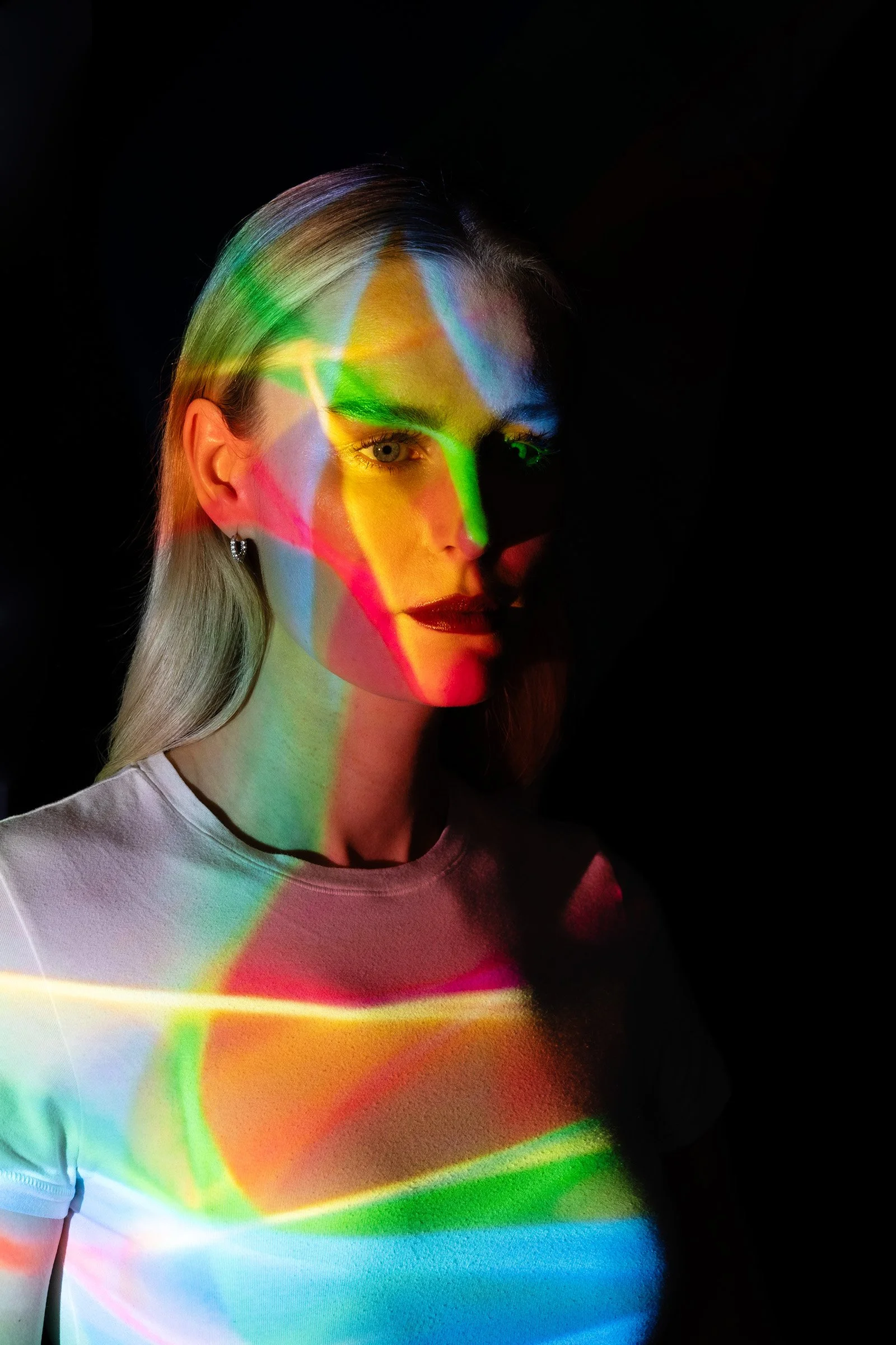 Junge Frau mit bunten Lichtreflexen auf Gesicht und Oberteil, dunkler Hintergrund.