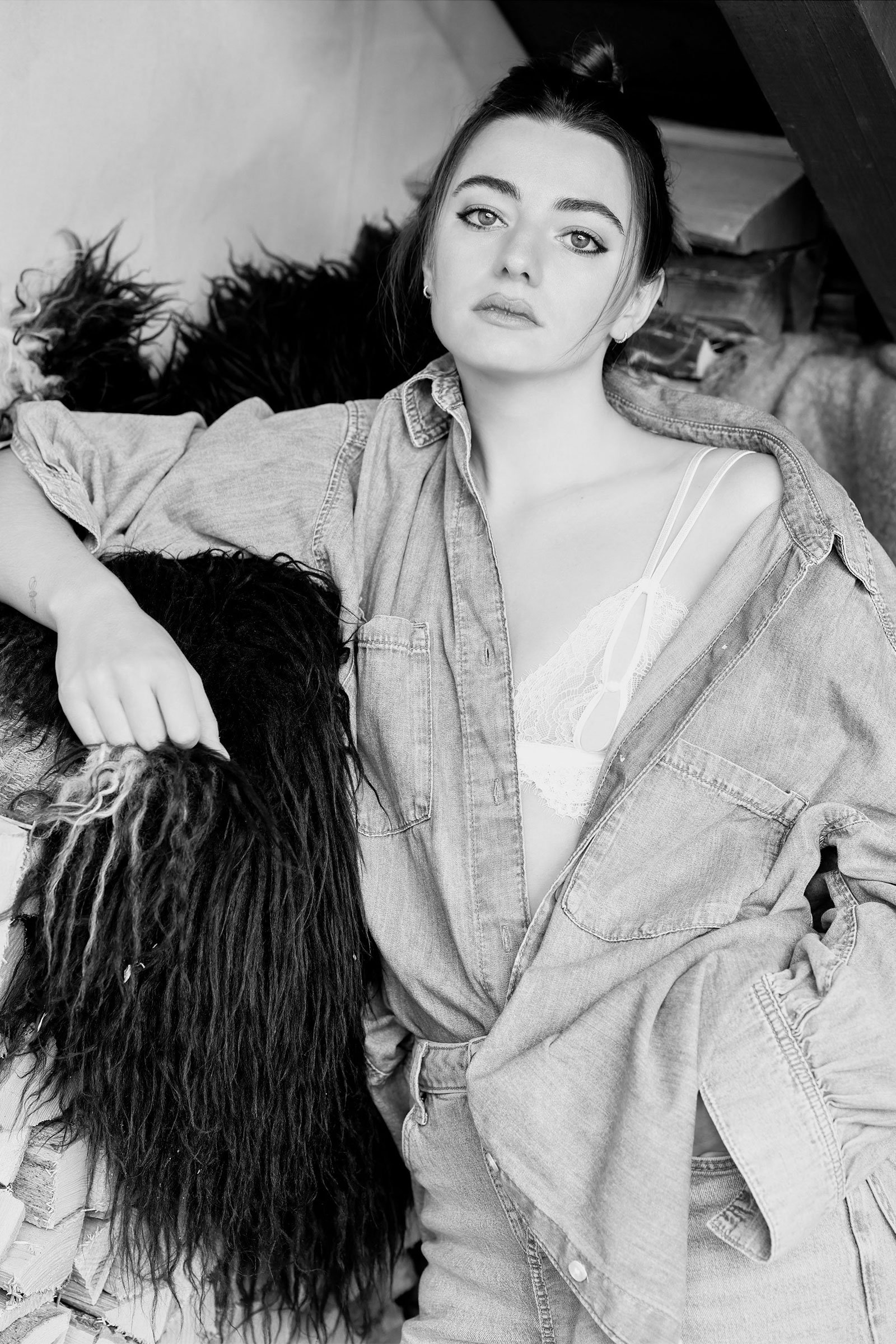 Schwarz-Weiß-Foto eines jungen Mädchens, das in Jeansjacke und Spitzen-BH sitzt, mit dunkler, lockiger Frisur, ruhigem Gesichtsausdruck, auf einem unstimmigen Hintergrund mit Holz und Fell.