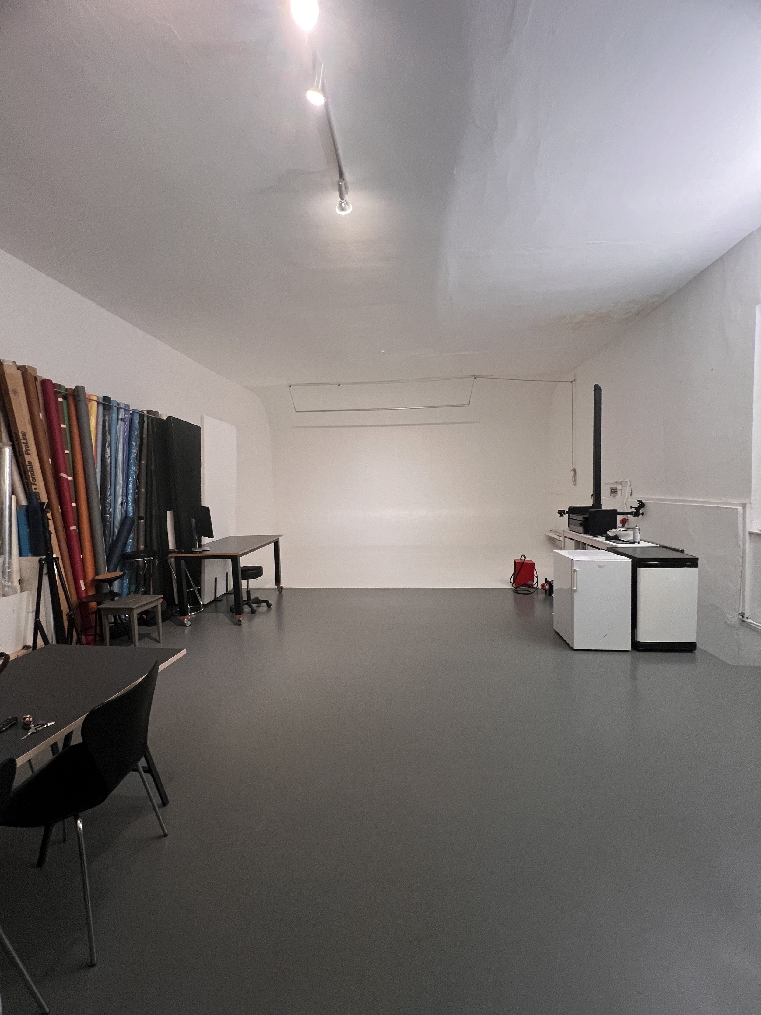 Leerer Raum mit weißen Wänden, grauem Boden, mehreren rollbaren Tischen, Stühlen und einer Staubschutzwand auf der linken Seite. Es gibt einige Büromaterialien, ein Messgerät und eine kleine Geräte auf der rechten Seite.