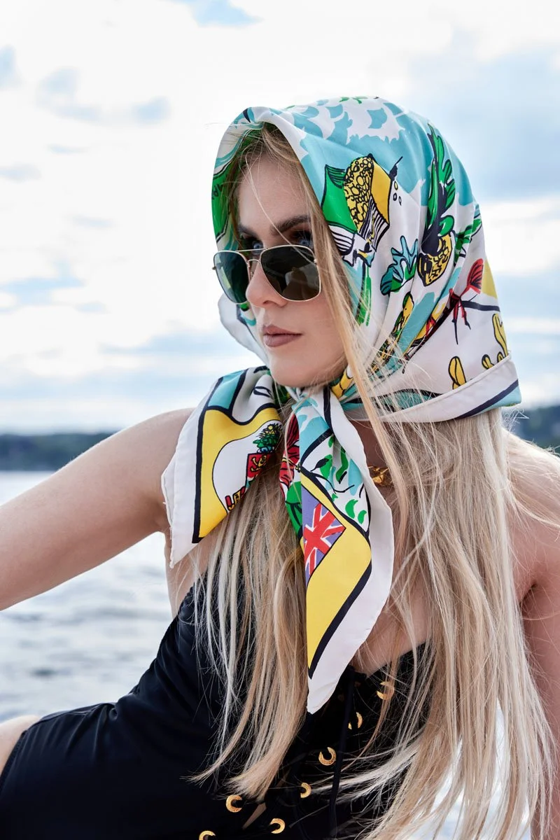 Eine blonde Frau trägt eine bunte Halstuch mit Tier- und Pflanzenmotiven und Sonnenbrille, sitzt bei Sonnenlicht am Wasser.