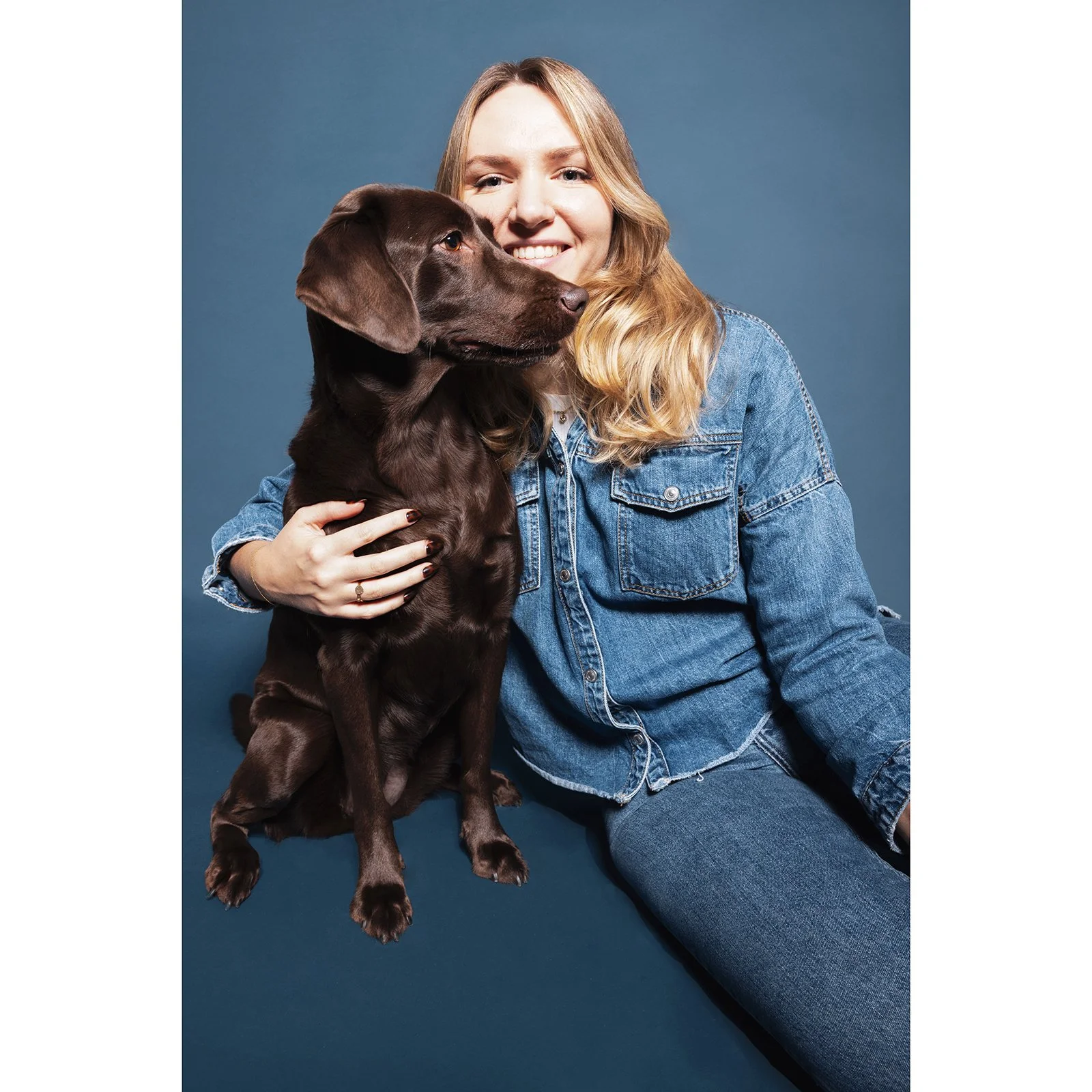Eine junge Frau in einem Jeanshemd hält einen braunen Hund auf dem Schoß, beide posieren vor blauem Hintergrund.