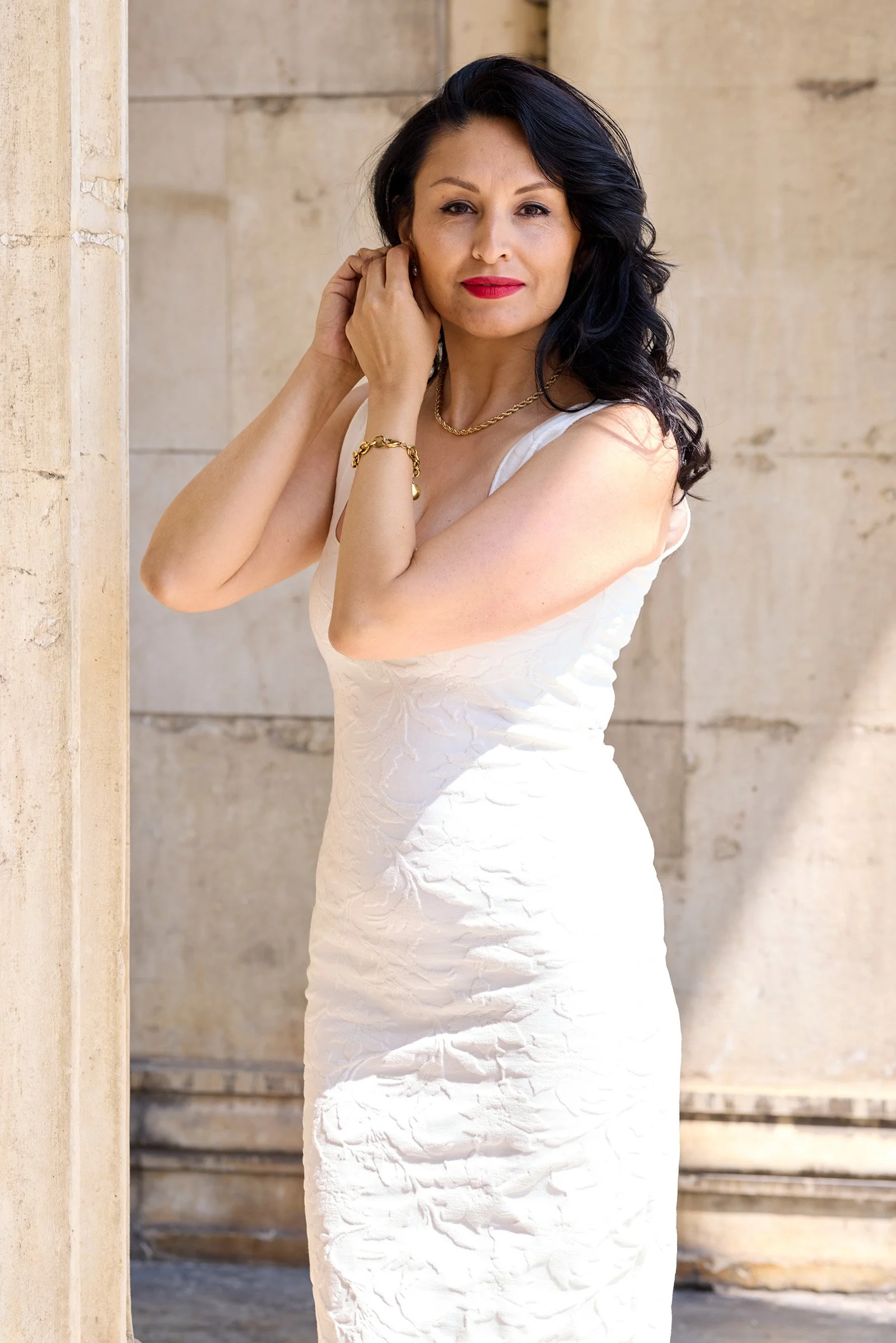 Frau in weißem Kleid mit dunklen Locken, rote Lippen, Goldschmuck, vor einer Steinfassade