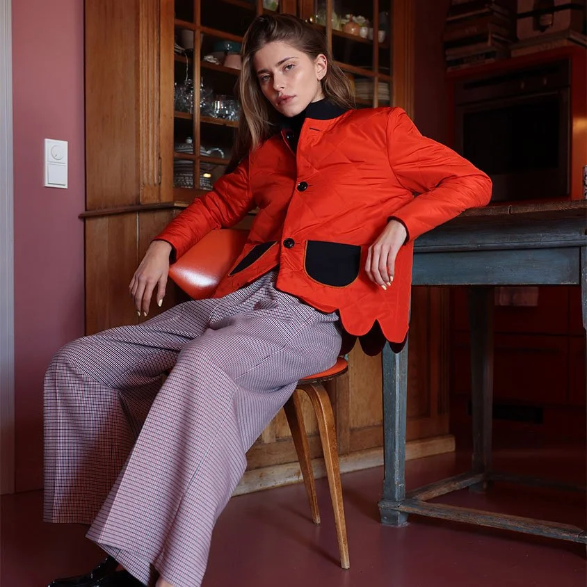 Eine junge Moderne Fashion affine Frau sitzt entspannt auf einem Stuhl in einem rustikalen Raum mit Holzmöbeln, trägt einen roten Blazer mit schwarzen Taschen und lila Karo-Hose.