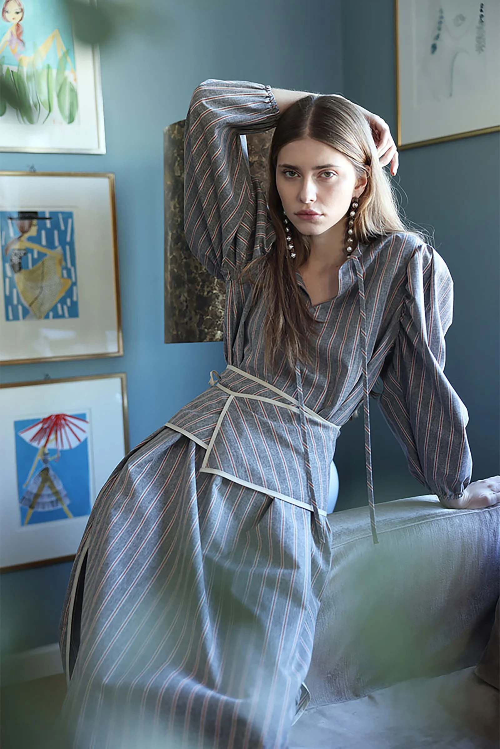 Junge Frau in gestreifter Bluse und Hose lehnt an Sofa, blickt ernst in die Kamera, im Hintergrund Kunstwerke an blauer Wand.