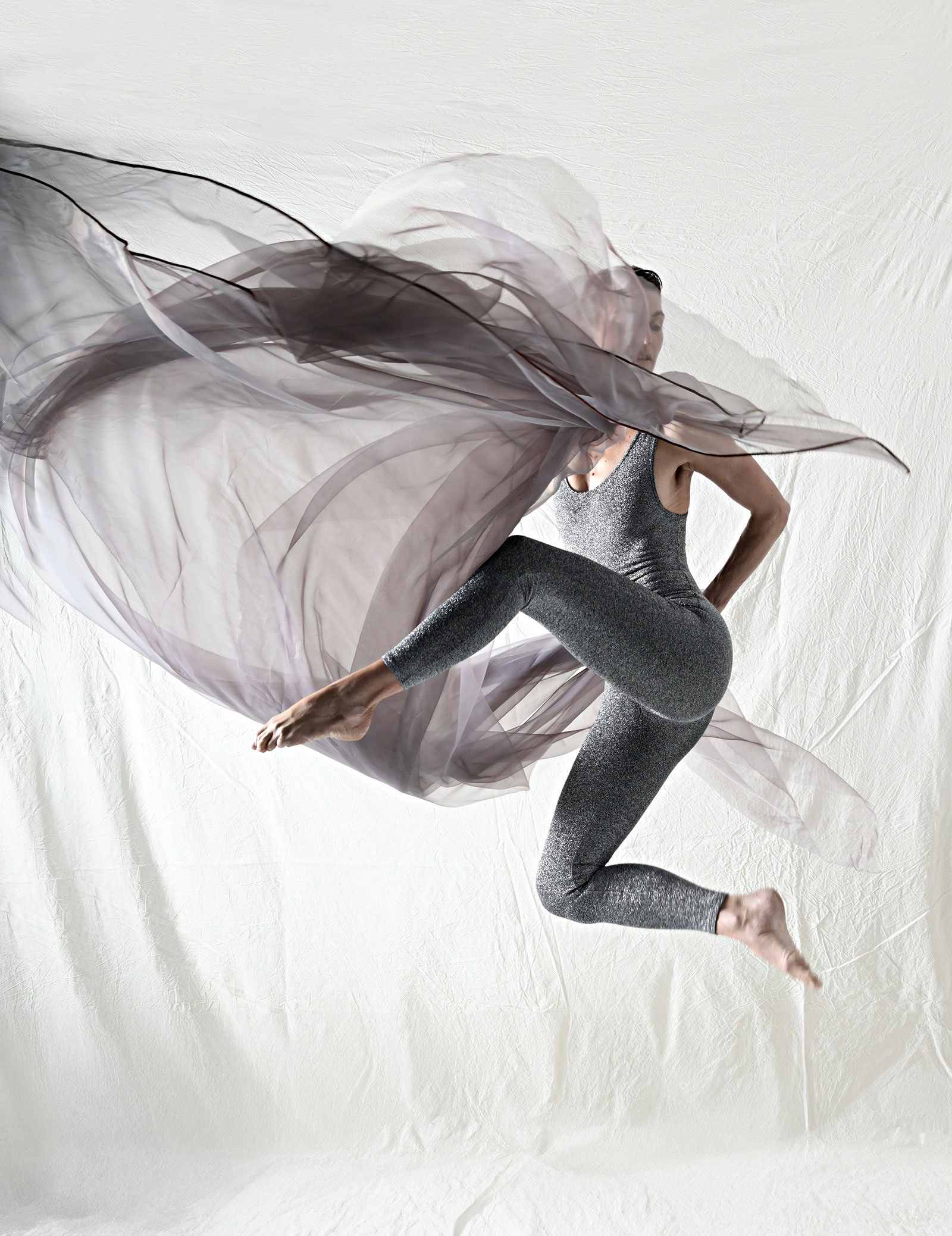 Tänzerin springt mit fließender Stoffoptik, trägt silbergrauen sportlichen Anzug, vor weißem Hintergrund.