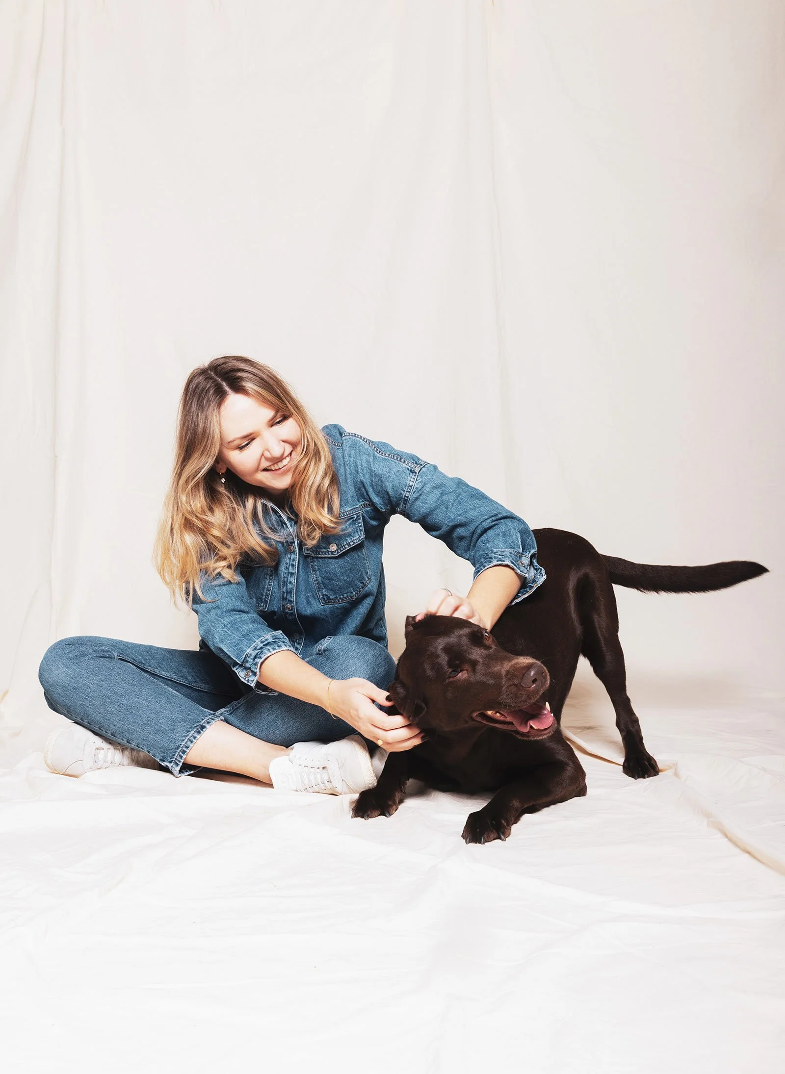 Eine Frau im blauen Jeans-Outfit sitzt lachend auf dem Boden und spielt mit einem braunen Labrador-Hund vor einem weißen Hintergrund.