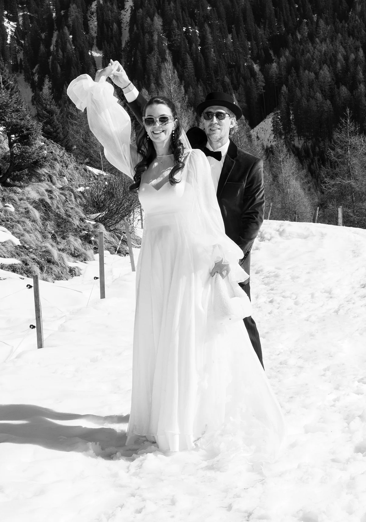 Ein Brautpaar, das im Schnee in den Bergen posiert, die Braut trägt ein langes weißes Kleid und der Bräutigam einen schwarzen Anzug mit Fliege und Hut, beide tragen Sonnenbrillen.