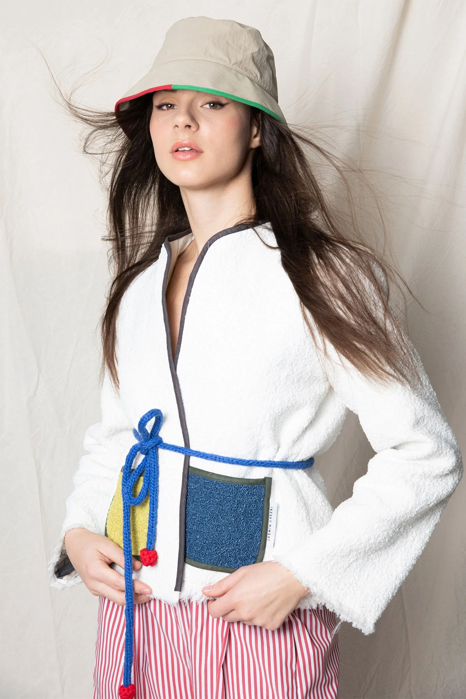 Frau mit langem braunem Haar trägt Hut, weiße Jacke mit Logo, rote und weiße gestreifte Hosen, und hat eine blaue Schnur um den Bauch sowie eine Tasche mit buntem Muster.