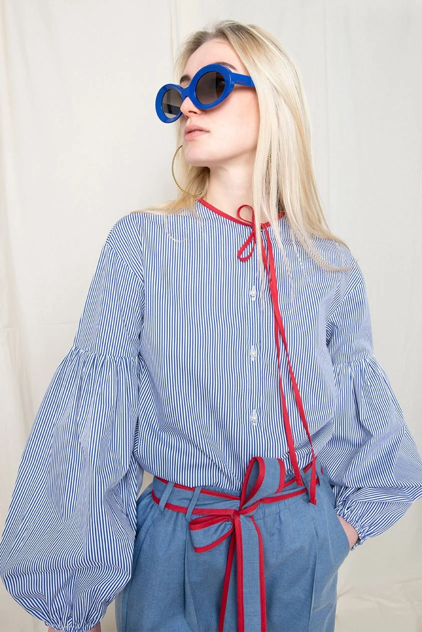 Frau mit blonden Haaren trägt große blaue Sonnenbrille, ein goldene Ohrring und ein blau-weiß gestreiftes Hemd mit rotem Detail an Ausschnitt und Bündchen.