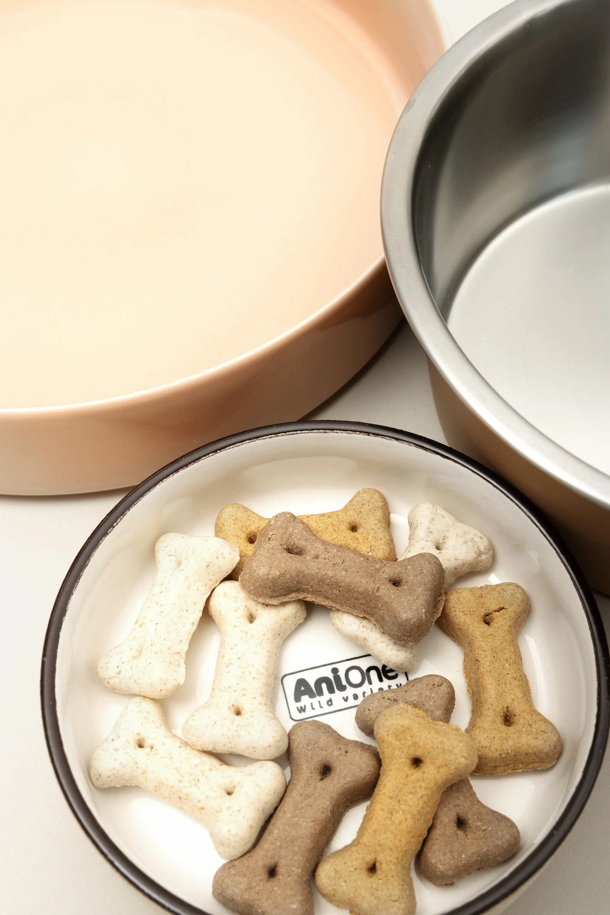 Eine Schüssel mit Knochen-förmigen Keksen in verschiedenen Farben mit dem Label 'AniOne Wild Variety', umgeben von zwei weiteren Schüsseln, eine beige und eine silberne.