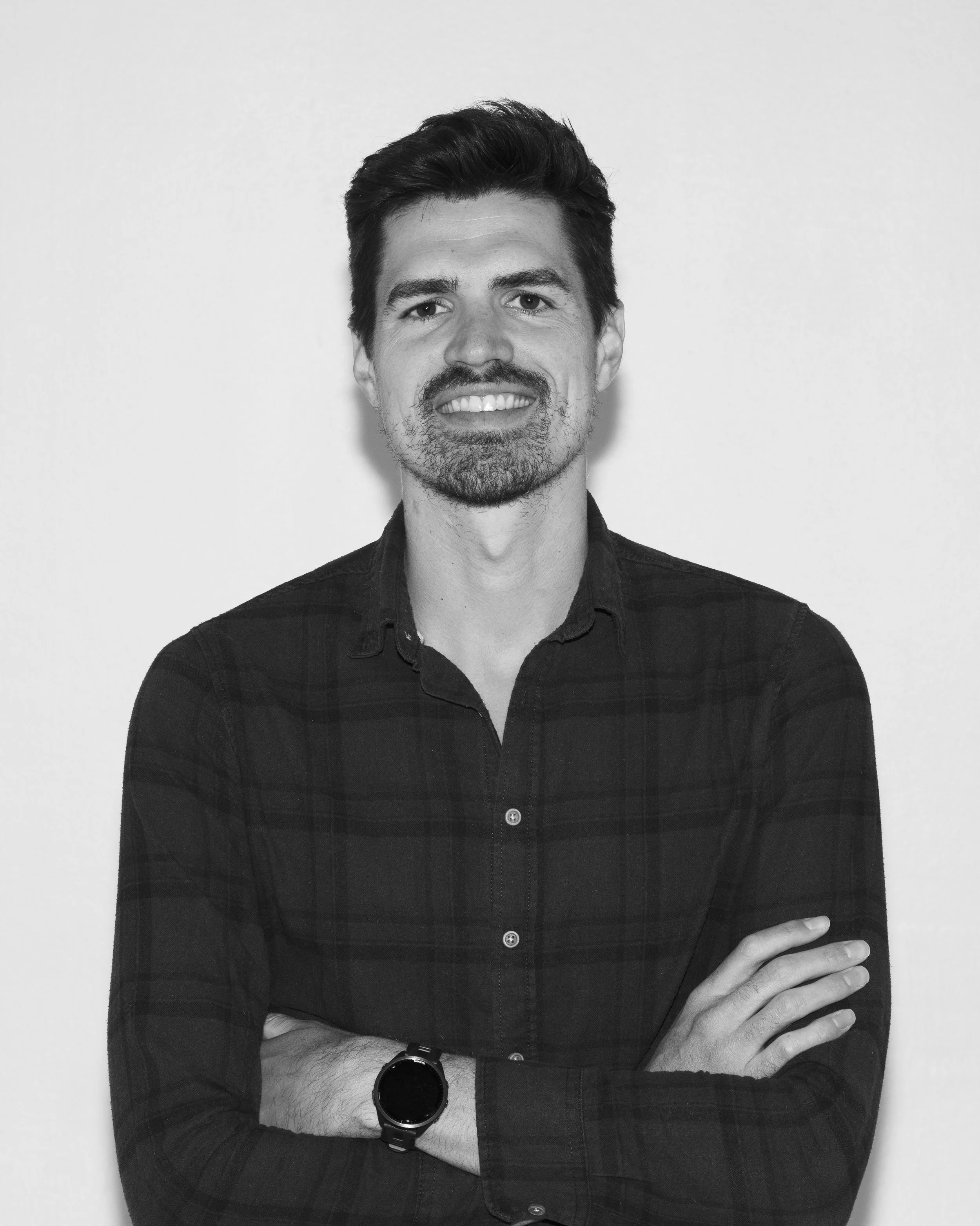 Schwarz-weiß Porträt eines Mannes, der lächelnd mit verschränkten Armen steht, trägt eine dunkle, kariert Hemd und eine Armbanduhr.