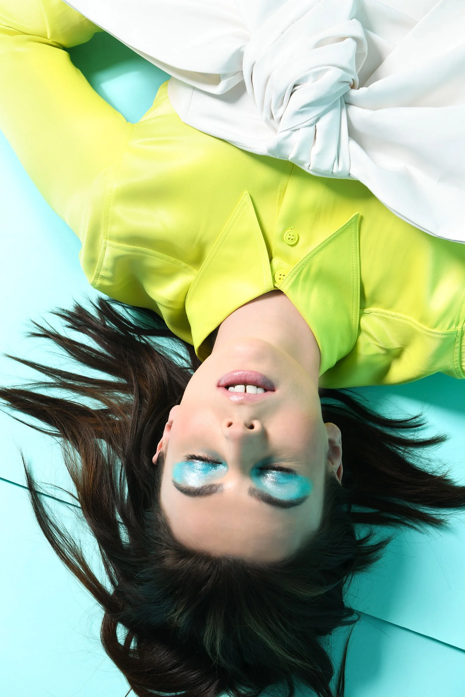 Eine Frau liegt auf dem Rücken auf einer blauen Oberfläche, trägt ein leuchtend gelbes Shirt mit großen, spitzen Kragen, hat lange braune Haare, die um sie herum liegen, und trägt blaue Lidschatten.
