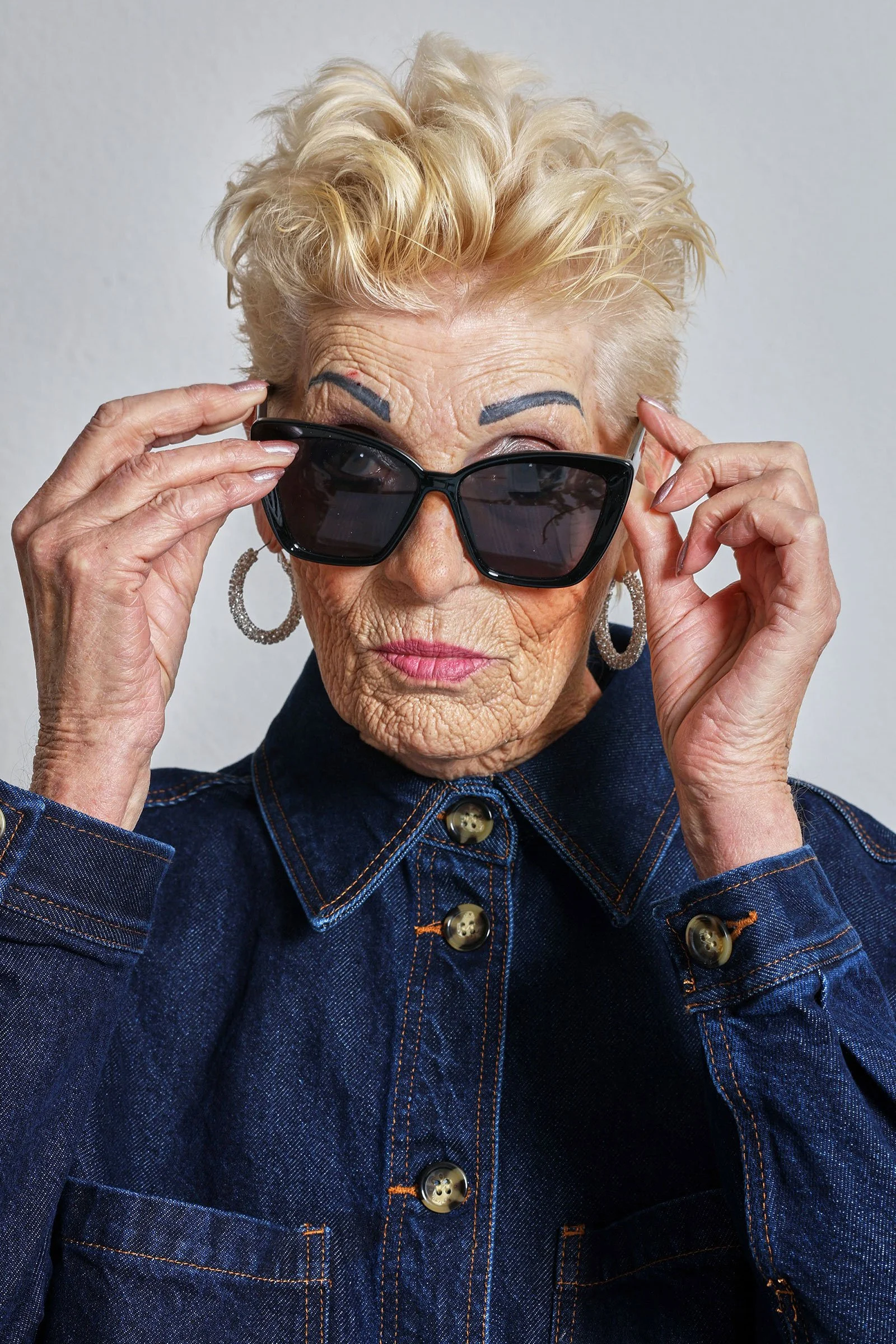 Ältere Frau mit blonden, lockigen Haaren trägt schwarze Sonnenbrille, große Ohrringe und eine dunkle Denim-Jacke. Sie sieht direkt in die Kamera und hebt die Sonnenbrille leicht an.