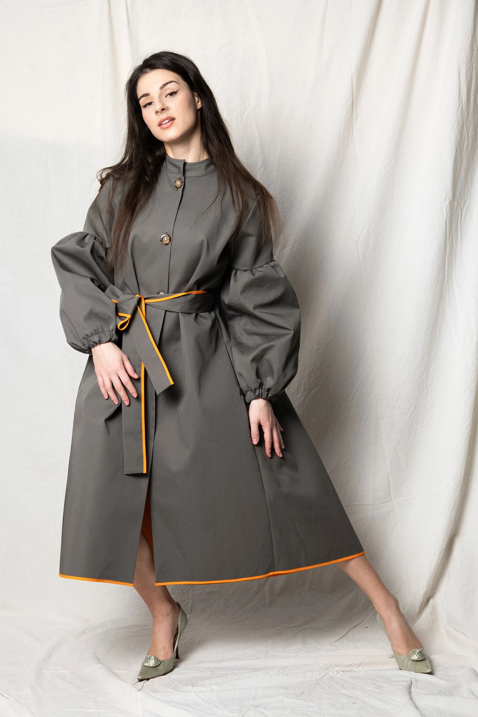 Frau in grauem Kleid mit orangefarbenem Saum und Gürtel steht vor weißem Hintergrund, posiert mit einem Bein nach vorne und einer Hand auf der Hüfte.