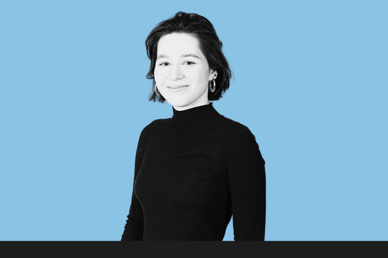 A woman with short dark hair, wearing a black turtleneck, smiling, against a light blue background.