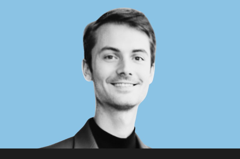 A young man with short dark hair, wearing a black turtleneck and blazer, smiling against a blue background.