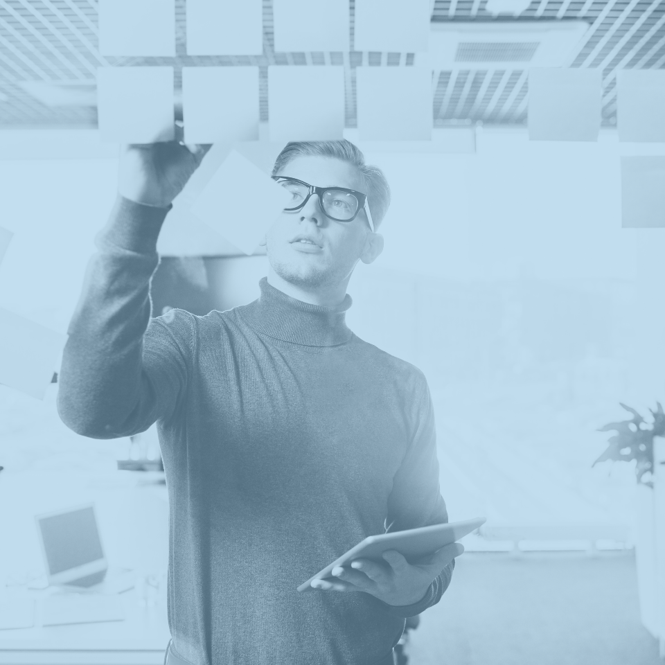 Man with glasses and a turtleneck writing notes on sticky notes attached to a ceiling grid, holding a tablet in his other hand.