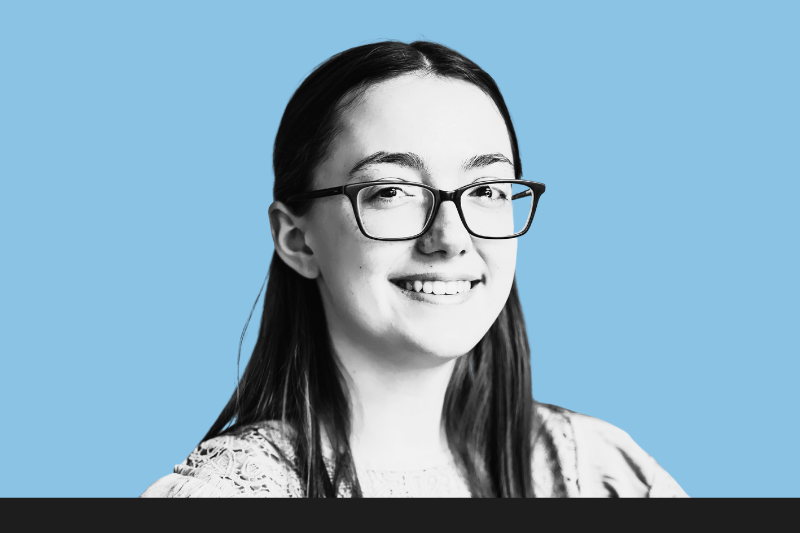 A young woman with long dark hair, wearing glasses and a lace top, smiling at the camera against a solid light blue background.