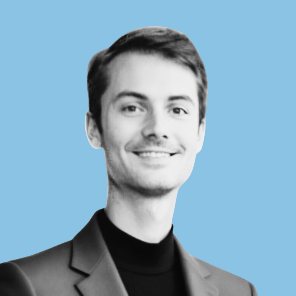 A young man with short dark hair, wearing a blazer and a black turtleneck, smiling against a light blue background.