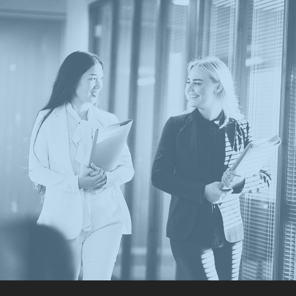 Two women in business attire walking and talking in an office hallway, holding folders and smiling.