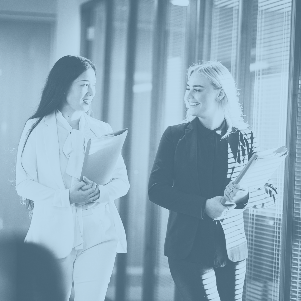 Two women in professional attire walking in an office hallway, carrying folders and smiling at each other.