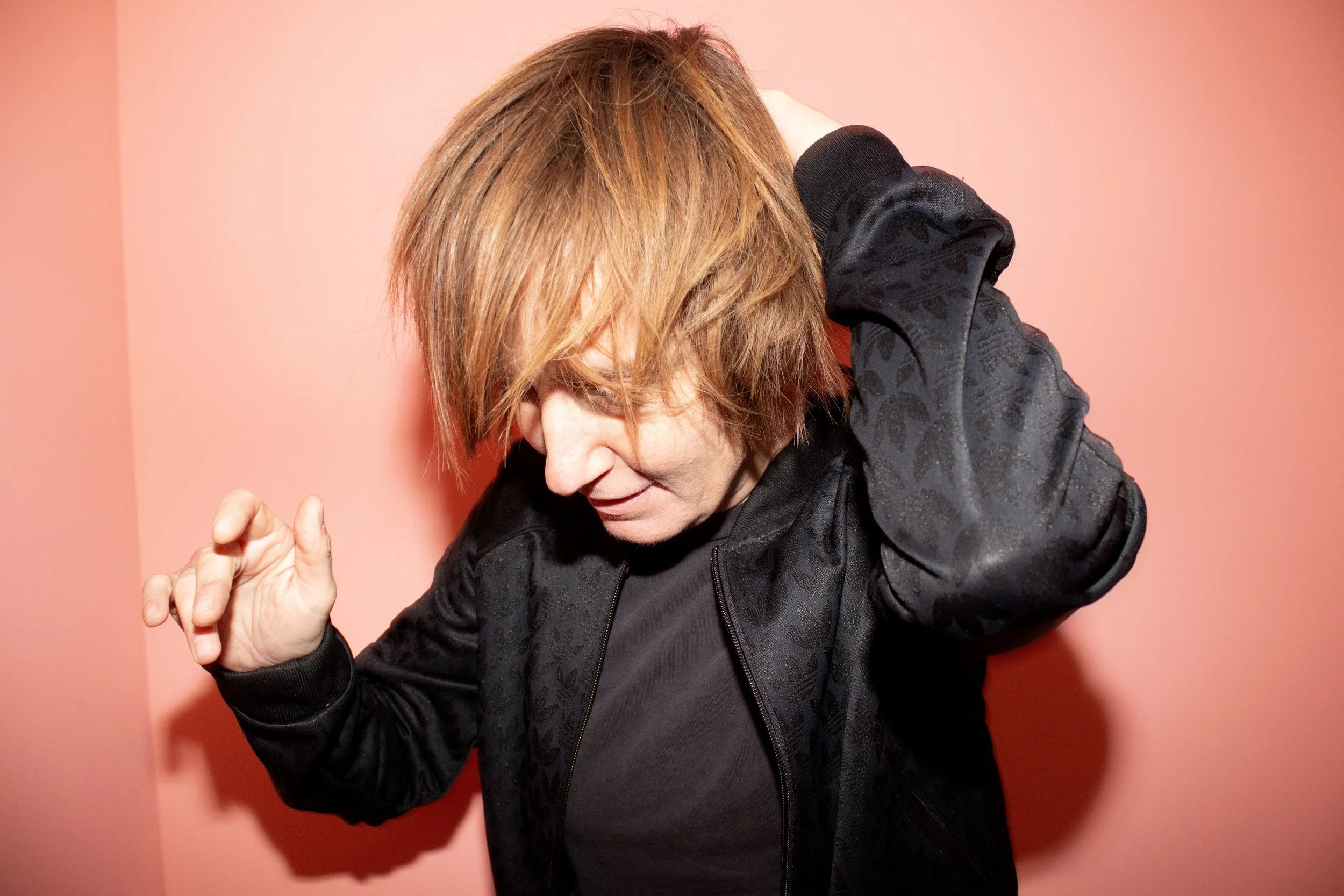 A woman with red hair wearing a black jacket, holding her head with one hand and making a gesture with her other hand, against a coral background.