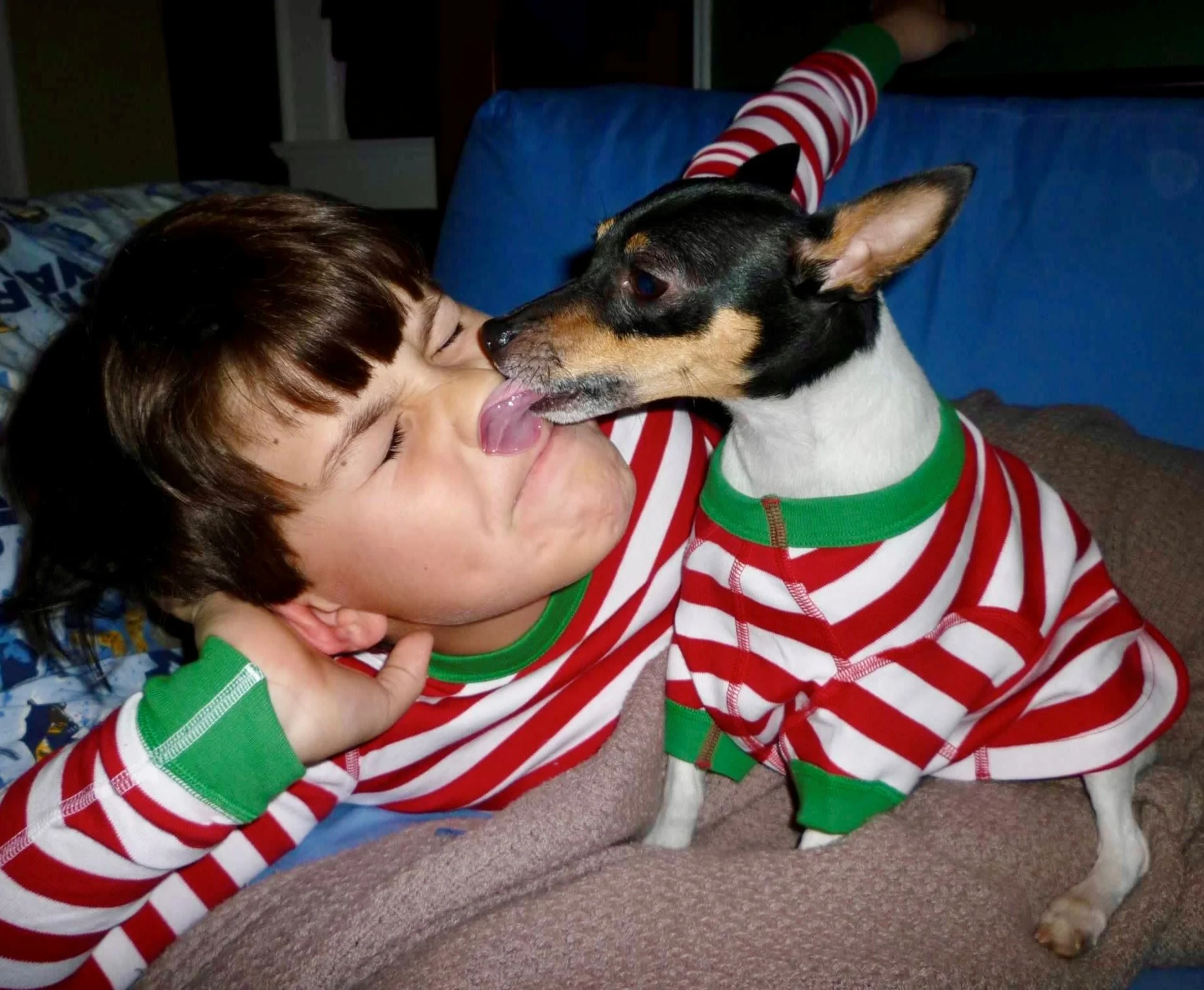 A young boy with brown hair and light skin lying on the floor, wearing a red and white striped shirt with green cuffs, being licked on the face by a small dog with black, white, and brown fur, wearing a matching red, white, and green striped shirt.