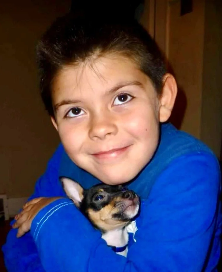 A boy in a blue shirt holding a small black and tan dog close to his shoulder.