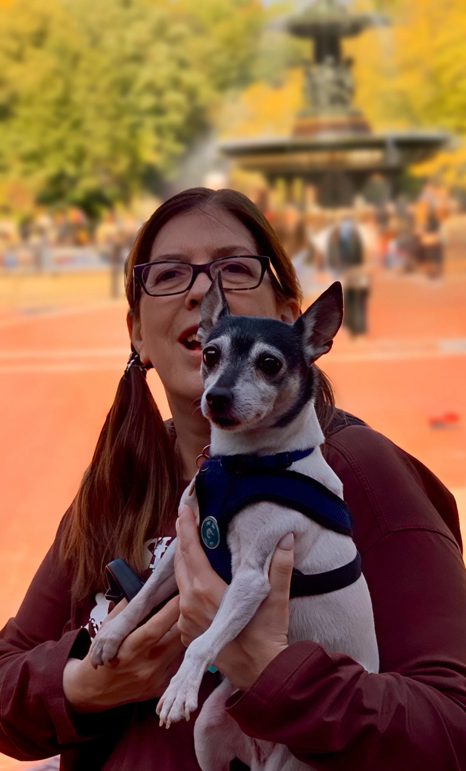 Woman with long hair and glasses holding a small black and white dog in front of a blurred park fountain with autumn trees.