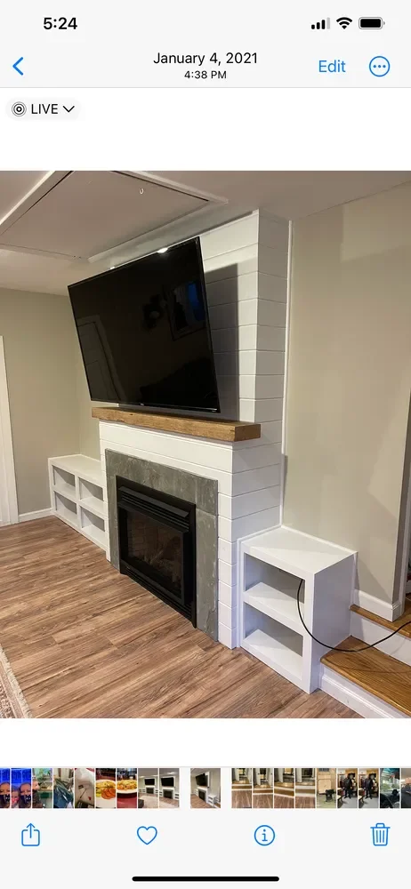 Living room fireplace with a mounted flat-screen TV above, white built-in shelves to the left, and a small white side table with an outlet cord to the right, on wood laminate flooring.