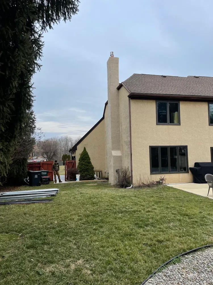 Backyard view of a beige two-story house with black window frames, a patio, and a large chimney on the side. A person stands near a red fence, and there is a large evergreen tree on the left. The lawn is green, and the sky is cloudy.