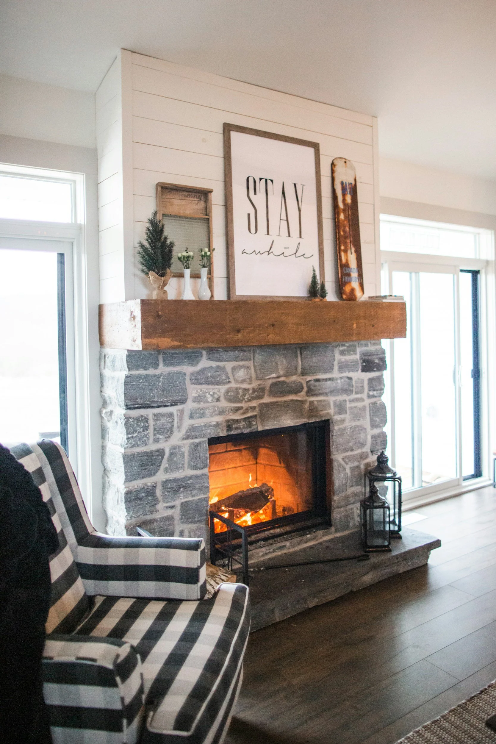 Living room with a stone fireplace, a plaid armchair, and farmhouse-style decor on the mantel, including small potted plants, a sign that says 'STAY awhile,' and a vintage skateboard, with large windows in the background.