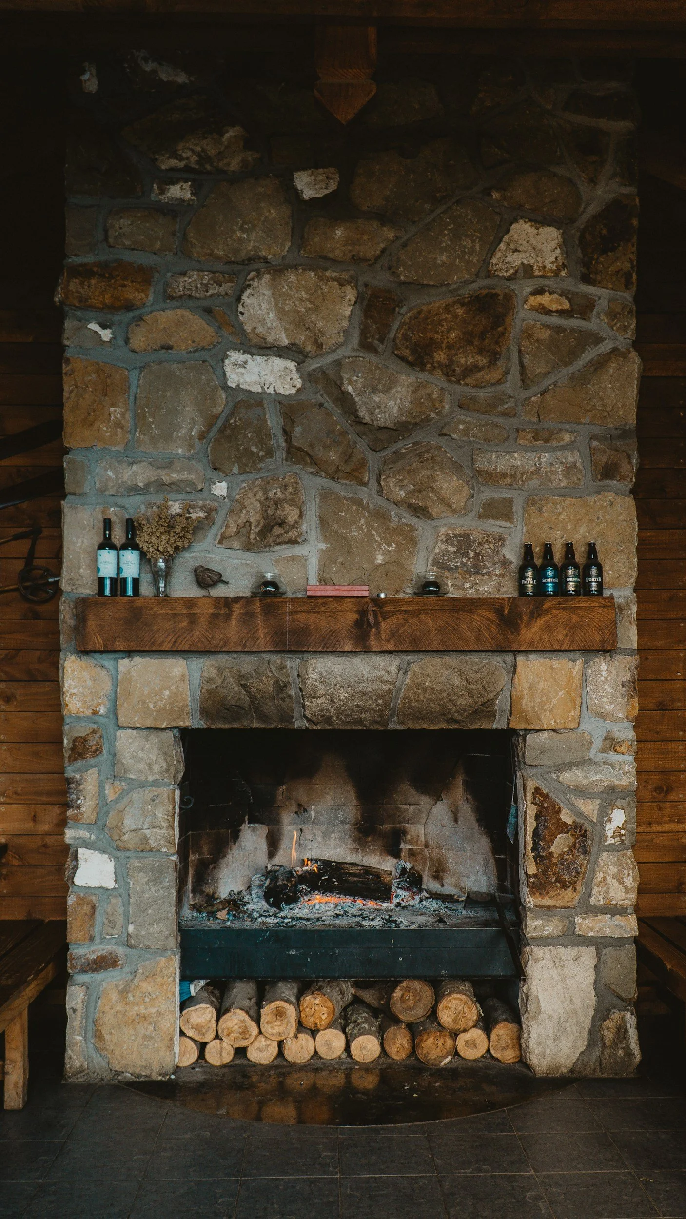 A large stone fireplace with a wooden mantel, some bottles, and decorative items on top. A fire is burning inside, with logs stacked underneath.