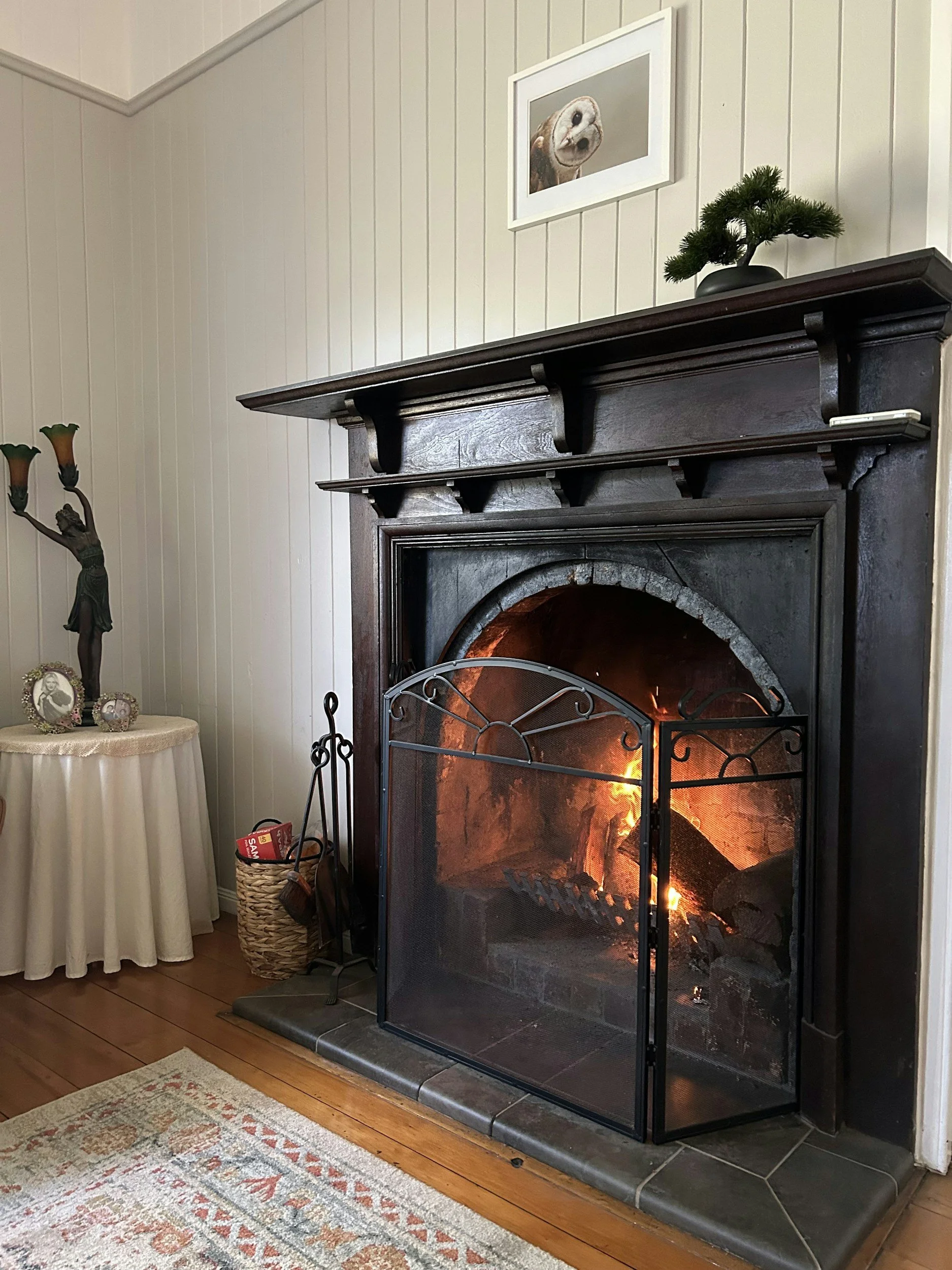 Living room with a lit fireplace, a small framed picture of a dog, a green plant on the mantle, a table with decorative items, and a basket with a fire tool set next to the fireplace.