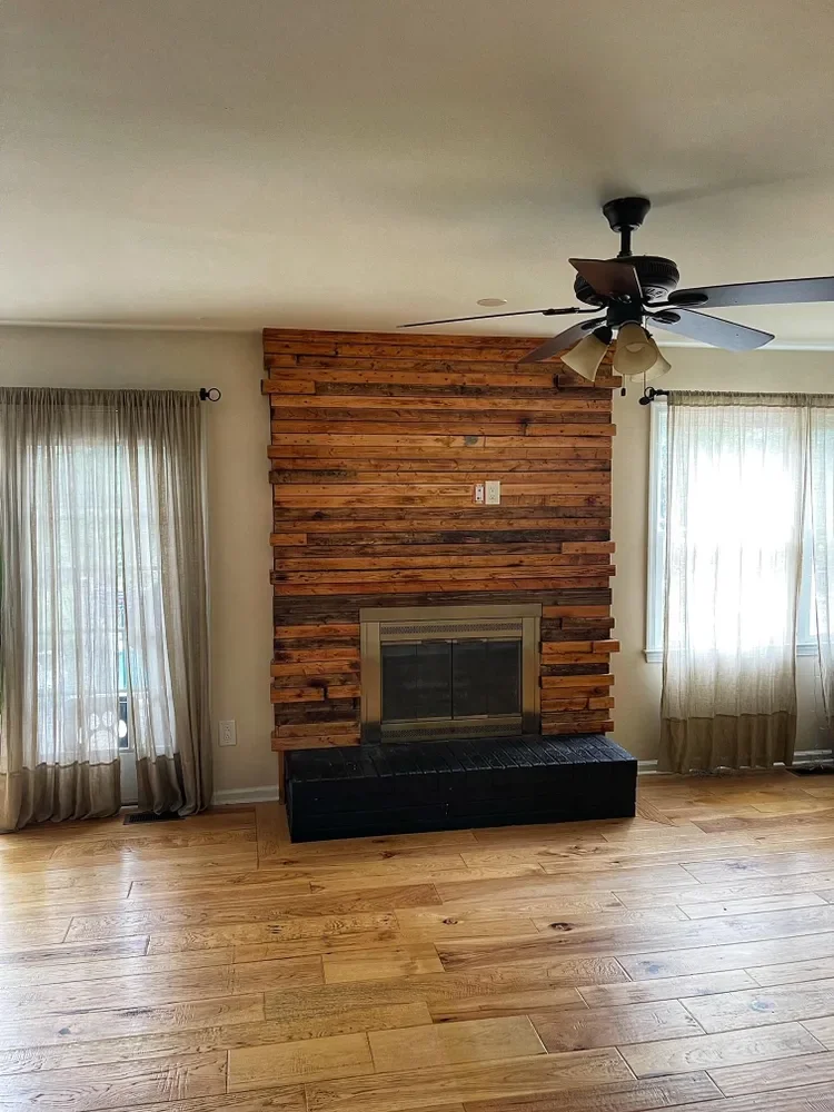 Living room with a wooden-paneled fireplace, beige curtains on windows, hardwood flooring, and a ceiling fan with light fixtures.