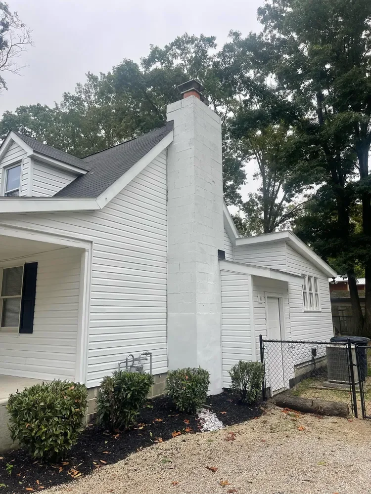 White house with a chimney, surrounded by trees and a small garden with bushes, black mulch, and a gravel walkway.