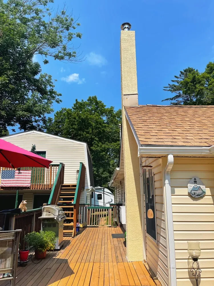 Backyard deck with wooden flooring, red umbrella, potted plants, a grill, a dog, and a neighboring house with a staircase leading up to a deck with an American flag, and trees under a blue sky.