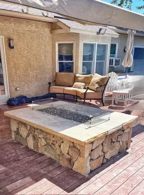 Outdoor patio with stone fire pit, beige sofa with pillows, white Adirondack chairs, and an umbrella in front of a beige house with windows.