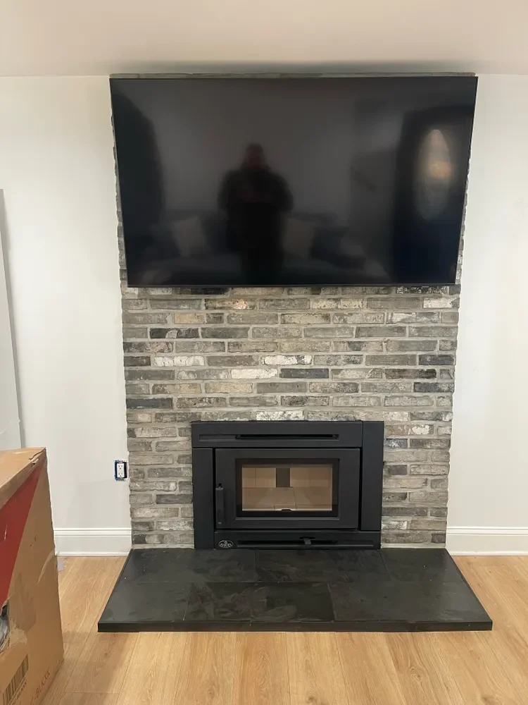 A flat-screen TV mounted above a brick fireplace with a black metal frame and glass door, sitting on a slate hearth on a wooden floor.