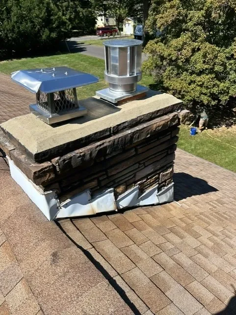View of a chimney with two metal vents on a house roof, side view showing shingles and a tree in the background.