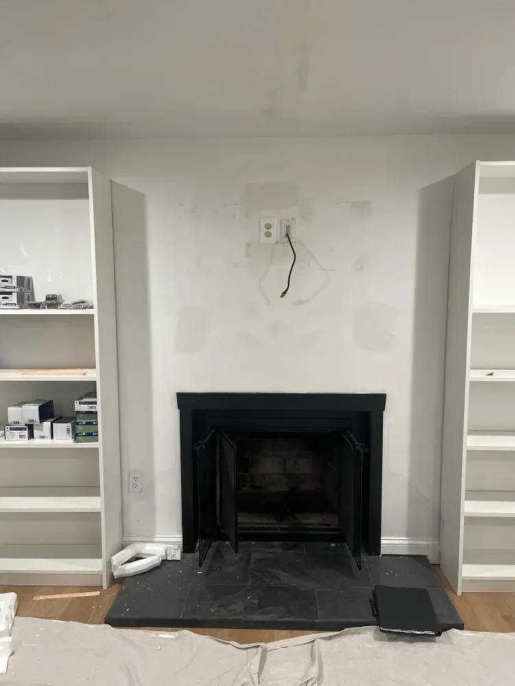 Living room fireplace with black metal frame, empty white built-in bookshelves on both sides, removed items, and an electrical outlet above the fireplace, with some construction materials and a cloth on the floor.