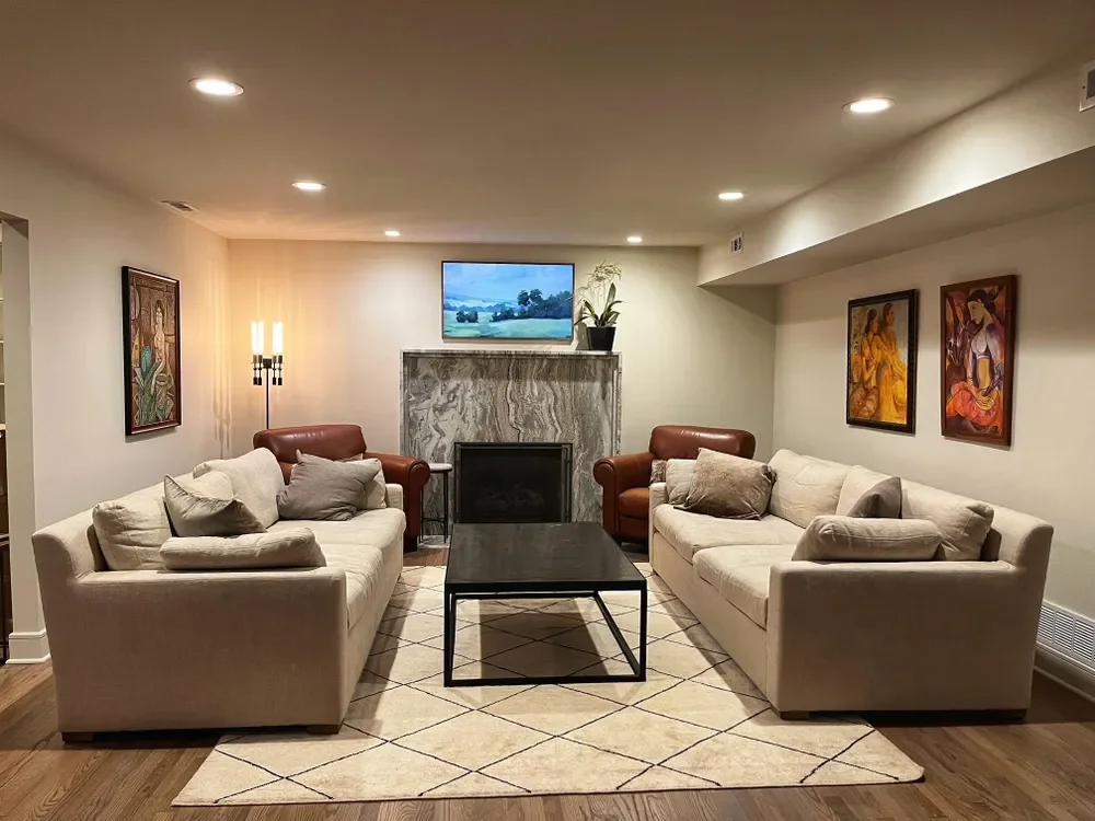 A cozy living room with two white sofas, two brown armchairs, a black coffee table, a fireplace with a marble surround, a mounted TV above the fireplace, artwork on the walls, and a beige rug covering a hardwood floor.