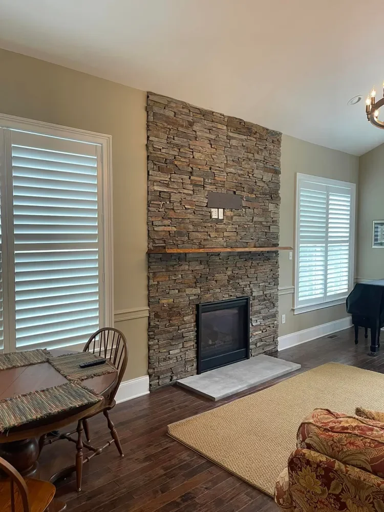 Living room with a stone fireplace, wooden floor, beige walls, windows with white shutters, a round dining table with chairs, a beige rug, and a floral armchair.