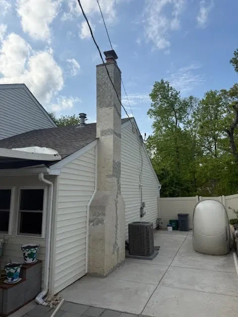 Backyard with a house, chimney, air conditioning unit, and an outdoor water heater or storage tank, with trees and a partly cloudy sky in the background.