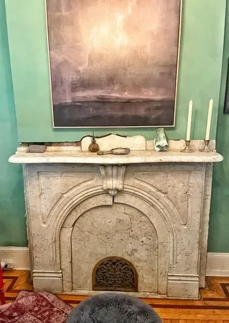 A vintage marble fireplace with decorative carvings, topped with a white marble mantle. On the mantle are two white candles in candleholders, a small green glass bottle, and small decorative items. A large abstract painting hangs above it, and the wall is painted green. Part of a patterned rug and a gray object are visible in the foreground.
