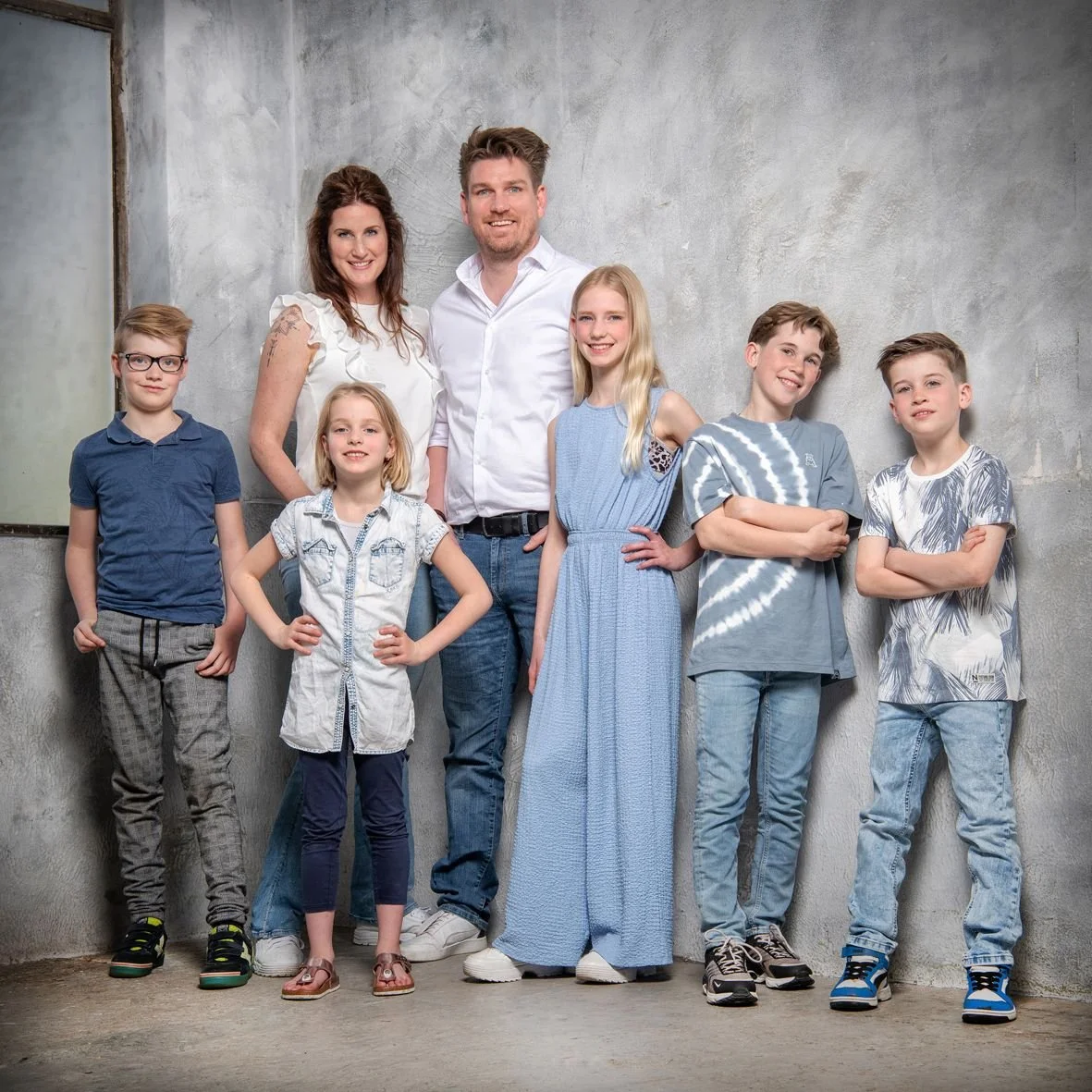 Gezinsfoto met twee volwassenen en vijf kinderen in een interieur met een betonnen muur.