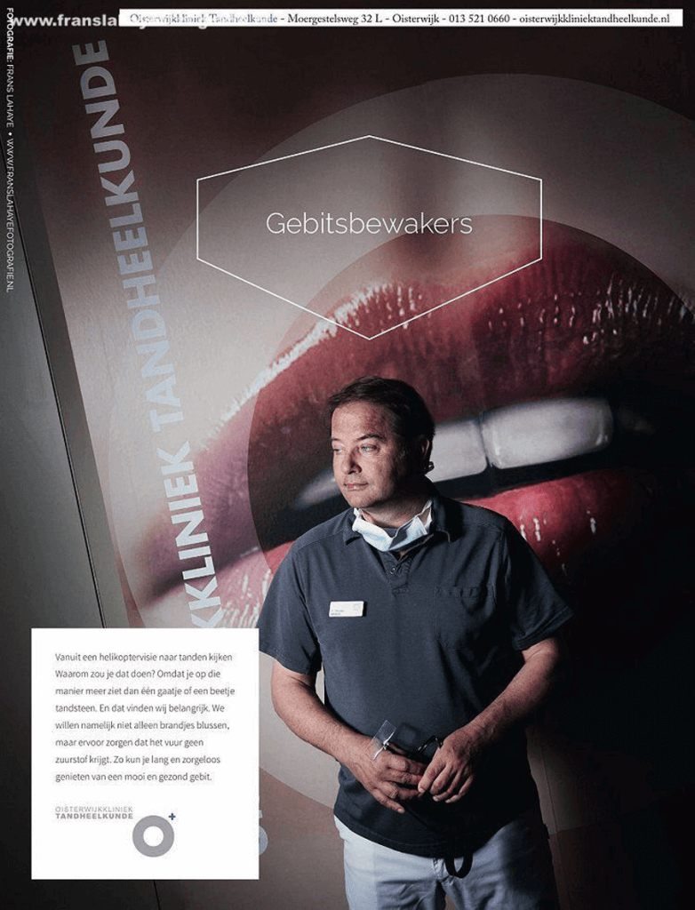 Een man in een uniform met een naamplaatje staat in een wachtkamer voor een mondgezondheid kliniek, met een poster over tandheelkunde achter hem. De poster bevat een grote afbeelding van lippen en tanden en de woorden 'Gebitsbewakers'.