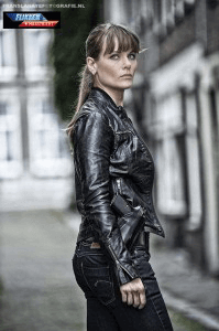 Vrouw in een zwarte leren jas staat op een straat in een stedelijk gebied, met een betonnen gebouw op de achtergrond.