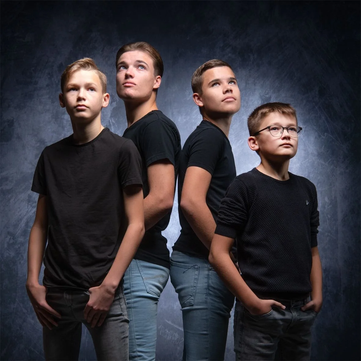 Vier jonge jongens in een donkere studio, gekleed in zwarte T-shirts en met verschillende haarkapsels, staan naast en achter elkaar met serieuze uitdrukkingen.