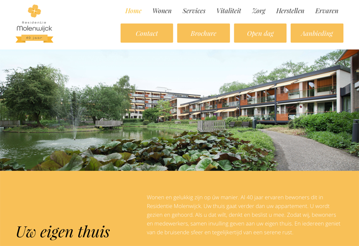 Een foto van een modern appartementencomplex aan een meer met een fontein, omringd door groene bomen en struiken, met paden en een bank in de tuin.